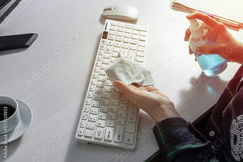 A girl in a checked shirt splashes cleaning agent on a rag to clean the keyboard. A Cup of coffee, a mouse, and a Notepad with a pen on a white office Desk. The concept of cleaning of the office