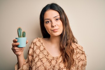 Wall Mural - Young beautiful brunette woman holding cactus pot over isolated white background with a confident expression on smart face thinking serious