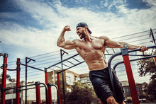 Bearded Bodybuilder Man Exercising On Monkey Bars For The Upper-body In A Modern Calisthenics Park Outdoors On A Sunny Day. Show Biceps And Scream For Motivation.