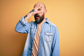 Poster - Handsome bald man with beard wearing casual denim jacket and striped t-shirt peeking in shock covering face and eyes with hand, looking through fingers with embarrassed expression.