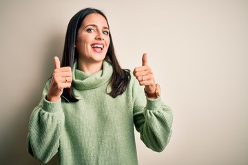 Young brunette woman with blue eyes wearing turtleneck sweater over white background success sign doing positive gesture with hand, thumbs up smiling and happy. Cheerful expression and winner gesture.