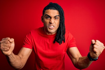 Wall Mural - Young handsome african american afro man with dreadlocks wearing red casual t-shirt angry and mad raising fists frustrated and furious while shouting with anger. Rage and aggressive concept.