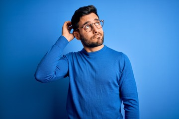 young handsome man with beard wearing casual sweater and glasses over blue background confuse and wo