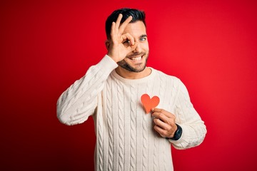 Wall Mural - Young handsome man holding red shape heart as romantic and health symbol with happy face smiling doing ok sign with hand on eye looking through fingers