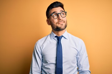 Young handsome businessman wearing tie and glasses standing over yellow background smiling looking to the side and staring away thinking.