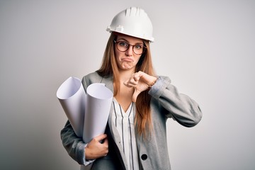 Wall Mural - Young beautiful redhead architect woman wearing glasses and safety helmet holding blueprints with angry face, negative sign showing dislike with thumbs down, rejection concept