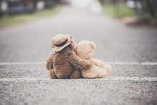 One Teddy Bear With His/her Arm Wrapped Around A Smaller Teddy Bear Showing Compassion On A Paved Street