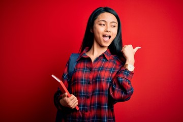 Wall Mural - Young beautiful chinese student woman holding book standing over isolated pink background pointing and showing with thumb up to the side with happy face smiling