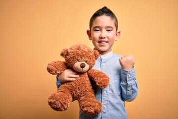 Poster - Young little boy kid hugging teddy bear stuffed animal over yellow background screaming proud and celebrating victory and success very excited, cheering emotion