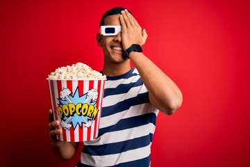 Young handsome african american man watching movie using 3d glasses eating popcorns covering one eye with hand, confident smile on face and surprise emotion.