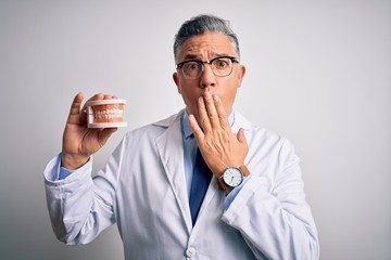 Poster - Middle age handsome grey-haired dentist man holding plastic teeth over white background cover mouth with hand shocked with shame for mistake, expression of fear, scared in silence, secret concept