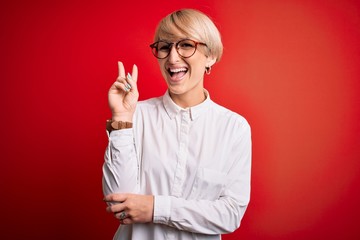 Wall Mural - Young blonde business woman with short hair wearing glasses over red background smiling with happy face winking at the camera doing victory sign. Number two.