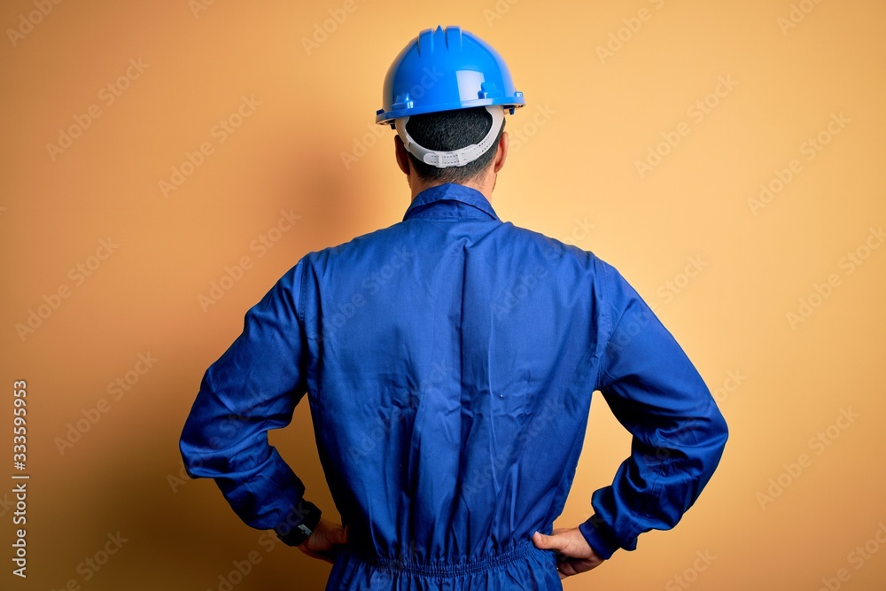 Mechanic man with beard wearing blue uniform and safety helmet over yellow background standing backwards looking away with arms on body - obrazy, fototapety, plakaty 