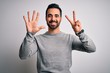 Young handsome man with beard wearing casual sweater standing over white background showing and pointing up with fingers number seven while smiling confident and happy.