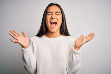 Young beautiful asian woman wearing casual sweater standing over white background celebrating mad and crazy for success with arms raised and closed eyes screaming excited. Winner concept