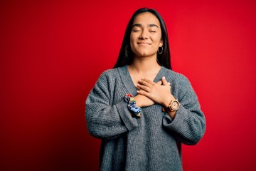 Canvas Print - Young beautiful asian woman wearing casual sweater standing over isolated red background smiling with hands on chest with closed eyes and grateful gesture on face. Health concept.