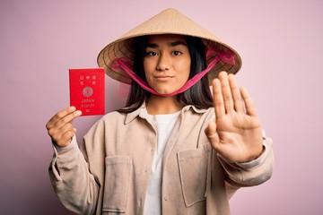 Wall Mural - Young thai woman wearing traditional conical asian hat holding japan japanese passport with open hand doing stop sign with serious and confident expression, defense gesture