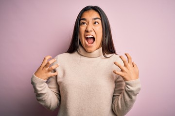 Young beautiful asian woman wearing casual turtleneck sweater over pink background crazy and mad shouting and yelling with aggressive expression and arms raised. Frustration concept.