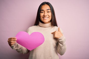 Canvas Print - Young beautiful asian woman holding pink heart standing over isolated background with surprise face pointing finger to himself