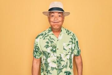 Sticker - Middle age senior grey-haired man wearing summer hat and floral shirt on beach vacation puffing cheeks with funny face. Mouth inflated with air, crazy expression.