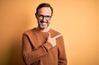 Middle age hoary man wearing brown sweater and glasses over isolated yellow background cheerful with a smile on face pointing with hand and finger up to the side with happy and natural expression