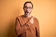 © Krakenimages.com - Middle age hoary man wearing brown sweater and glasses over isolated yellow background Surprised pointing with finger to the side, open mouth amazed expression.