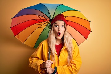 Sticker - Young beautiful blonde woman wearing raincoat and wool cap holding colorful umbrella scared in shock with a surprise face, afraid and excited with fear expression