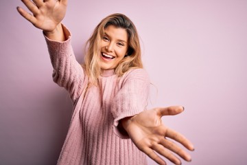 Canvas Print - Young beautiful blonde woman wearing casual pink sweater over isolated background looking at the camera smiling with open arms for hug. Cheerful expression embracing happiness.