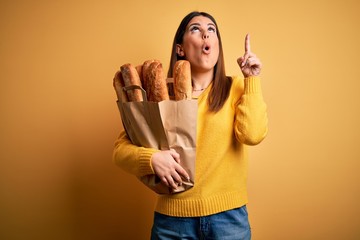 Poster - Young beautiful woman holding a bag of fresh healthy bread over yellow background amazed and surprised looking up and pointing with fingers and raised arms.