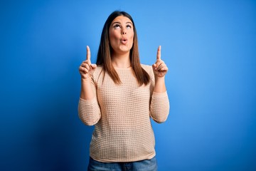 Sticker - Young beautiful woman wearing casual sweater over blue background amazed and surprised looking up and pointing with fingers and raised arms.