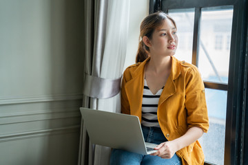 Asian beauty female sit next to window with light glow freelance work at home for protection covid-19 idea concept.Attractive asian woman happiness working with laptop in yellow casual dress at home.