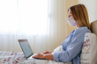 Close up shot of young blonde woman wearing disposable face mask working at home due to coronavirus quarantine concept. Beautiful female sitting in bed with laptop. Background, copy space.