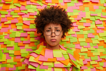 Wall Mural - Unhappy distressed Afro American woman thinks about something troublesome, wants to cry, feels bad and offended, being insulted, wears round eyewear. Head through wall with colored stickers.