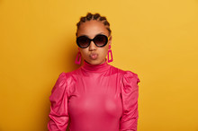 Beautiful Cheeky Woman Keeps Lips Rounded, Wears Trendy Big Sunglasses And Pink Blouse, Has Glamour Look, Isolated On Yellow Background. Fashion Model Dressed In Stylish Outfit. Beauty Trends