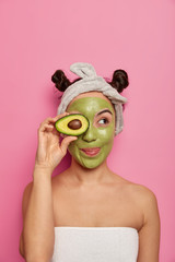 Studio shot of young female model feels fresh, holds avocado, applies green mask, cleans skin from pores, wears headband, has two knots, looks aside, isolated on pink background, copy space.