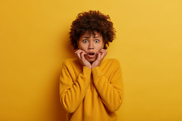 Wall Mural - Oh gosh, how scaryig! Emotional shocked woman grabs face and stares with surprise, realizes she lost everything, gasps from wonder, reacts amazed and speechless, wears casual jumper, yellow background