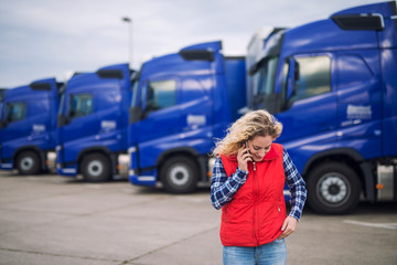 Female trucker talking on the phone about shipment that has to be delivered. Truck vehicles parked in background.