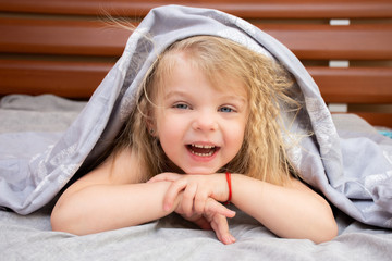 Portrait of a blue-eyed little girl with blond curly hair waking up in the morning on a gray bed laughing merrily.