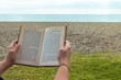 © Kemedo - Background of a person reading on the beach. Hands holding a book with the sea and sky in the background with copy space.