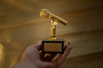 woman white hand holding gold, metallic microphone, mike, mic statuette, figurine medal award for mu