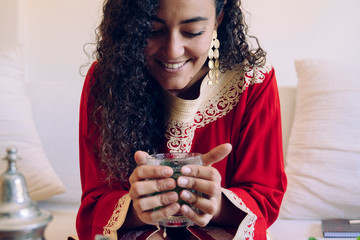 Beautiful young ethnic woman drinking herbal tea at home. Moroccan female with traditional clothes holding arabic beverage for healthy detox lifestyle. Morocco culture and traditions. Muslim concept.