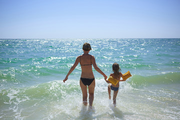 Wall Mural - Summer happy family of six years blonde child playing and jumping water waves embracing woman mother in sea shore beach