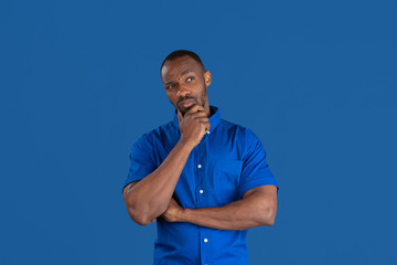 Wall Mural - Thinking, dreaming. Monochrome portrait of young african-american man isolated on blue studio background. Beautiful male model. Human emotions, facial expression, sales, ad concept. Youth culture.