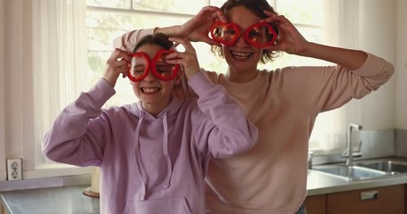 Wall Mural - Funny young mom and teenage daughter making pepper glasses having fun. Happy teen girl helping mum in kitchen laughing, looking at camera. Cheerful vegan family enjoying cooking activity, portrait.