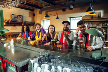 Wall Mural - Sport fans cheering at bar, pub and drinking beer while championship, competition is going. Multiethnic group of friends excited watching translation. Human emotions, expression, supporting concept.