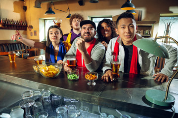 Wall Mural - Sport fans cheering at bar, pub and drinking beer while championship, competition is going. Multiethnic group of friends excited watching translation. Human emotions, expression, supporting concept.