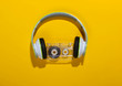 Stylish wireless stereo headphones and audio cassette on yellow background with shadow. Music lover. Retro 80s. Top view.