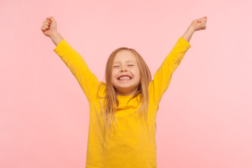 Child sincerely rejoicing success. Cute energetic enthusiastic little girl raising arms in excitement and keeping eyes closed, enjoying victory, winning. indoor studio shot isolated on pink background