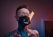 © Fxquadro - Bold and rebellious male model posing in a neon studio wearing a medical mask on his face, yellow sunglasses and tattooed in a japanese irezumi style of a half-naked body, looking self-assured.
