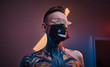© Fxquadro - Bold and rebellious male model posing in a neon studio wearing a medical mask on his face, yellow sunglasses and tattooed in a japanese irezumi style on a half-naked body, looking self-assured.